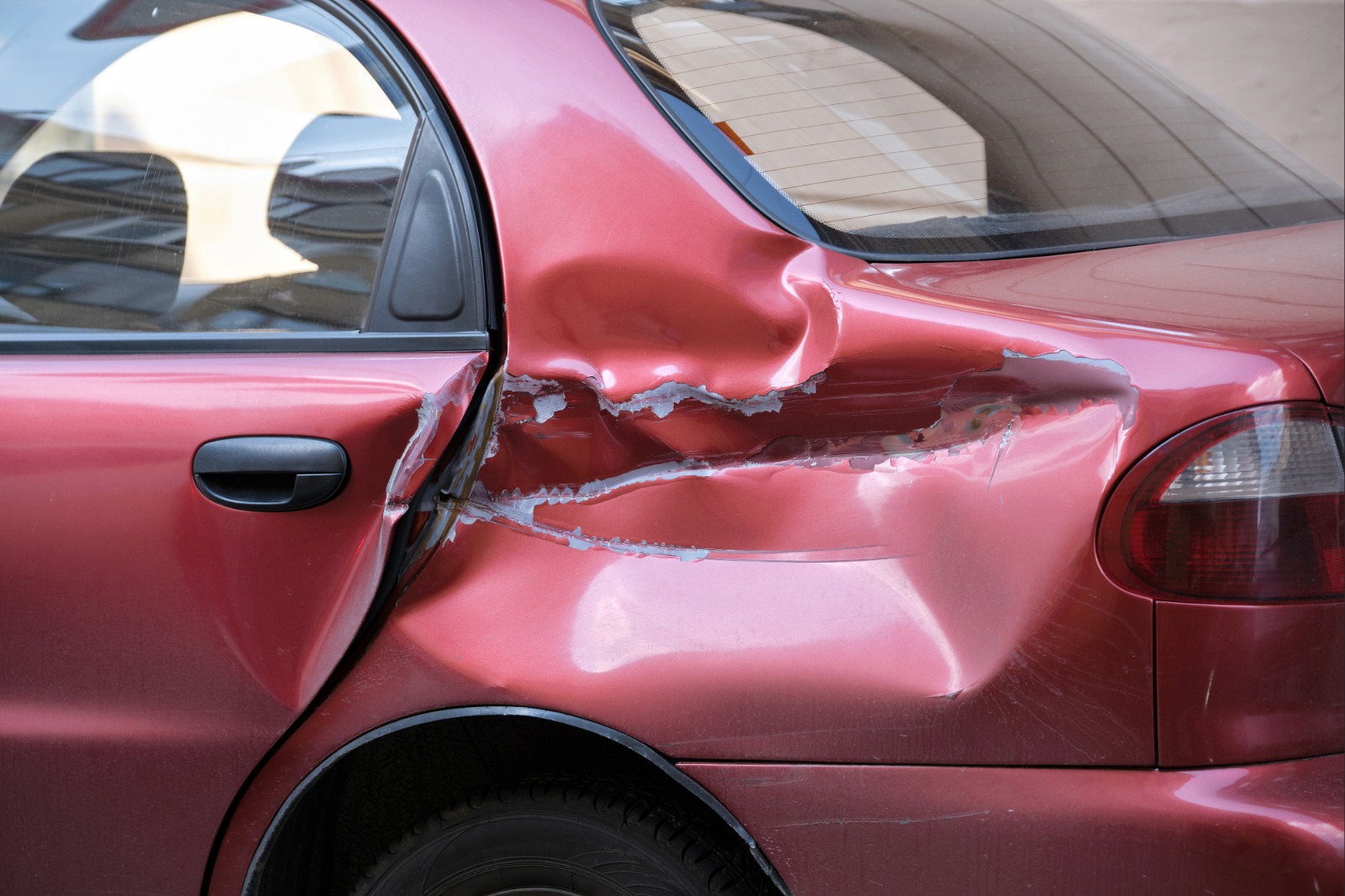 Stark besch&auml;digte Seitenpartie eines roten Autos mit tiefen Dellen, Kratzern und abgeplatztem Lack.